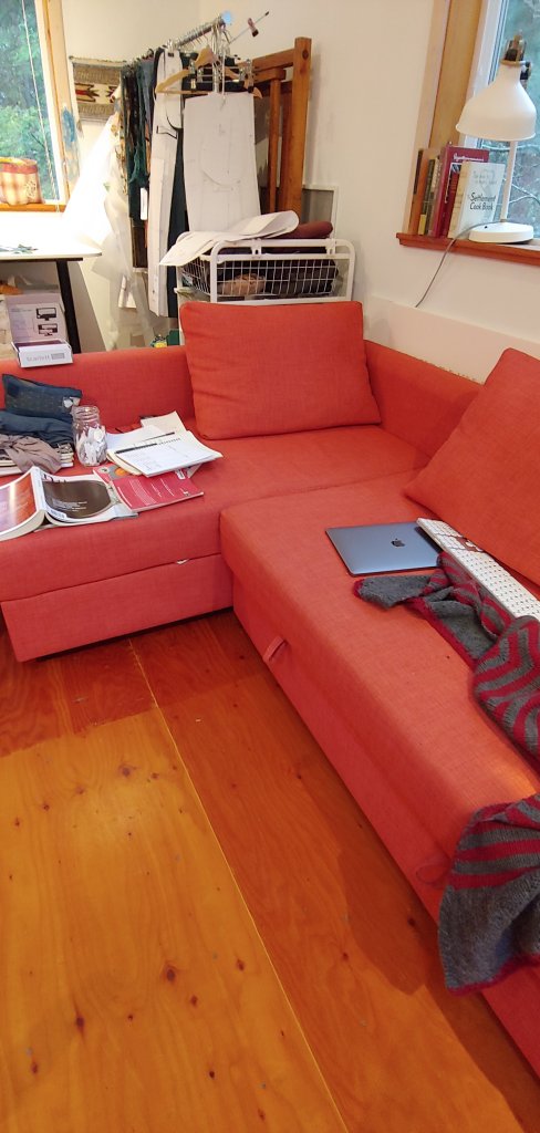 A photograph of a couch with books, papers and a laptop strewn around. 